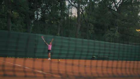 Eine-Frau-Im-Tennistraining-Trainiert-Einen-Schlagball-Und-Dann-Eine-Rückhand.-Outdoor-Training-Bei-Sonnenuntergang.-Ein-Sandplatz-Mit-Grünem-Zaun.-Weitwinkel-Durch-Das-Netz.